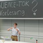 Ein Studi steht am Rednerpult mit dem Tafelaufschrieb "SCIENCE-TOK Vorlesungen?" auf der Tafel hinter ihm.