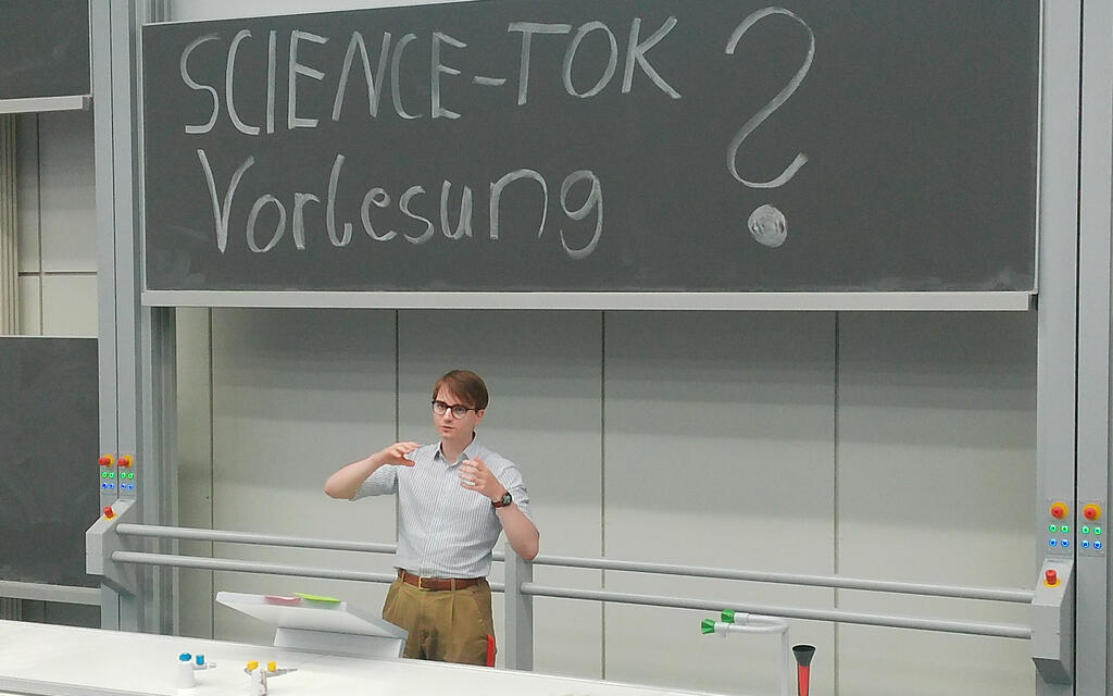 Ein Studi steht am Rednerpult mit dem Tafelaufschrieb "SCIENCE-TOK Vorlesungen?" auf der Tafel hinter ihm.